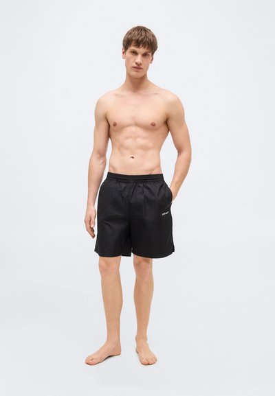 Jeune homme torse nu debout, pieds nus, portant un short noir au genou, une main dans la poche, sur un fond clair uni.