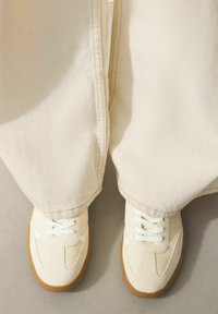 Baltos sportbačiai su perforuotais akcentais ir šviesiomis raištelėmis, derinami su laisvomis smėlio spalvos kelnėmis, matomi iš viršaus.