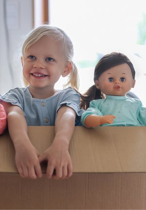 Uśmiechnięta młoda dziewczyna o blond włosach trzyma duże kartonowe pudełko zawierające lalkę z ciemnymi włosami i turkusowym strojem.