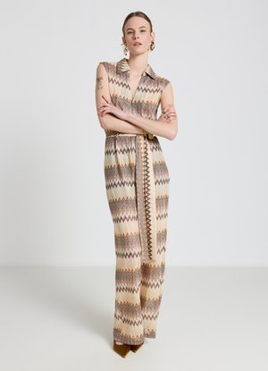 Donna in piedi con le braccia incrociate, indossa una tuta senza maniche beige e marrone con motivo a zigzag e scarpe marroni con punta su sfondo uniforme.