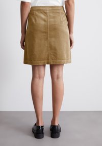 Mini-jupe en velours côtelé beige avec une coupe droite, dotée d'une taille et de deux poches arrière, associée à des chaussures noires. Texture lisse, motif à côtes.