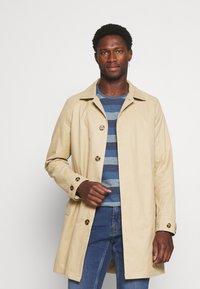 Trench coat beige realizzato in tessuto leggero, con design monopetto, bottoni grandi e colletto a punta.