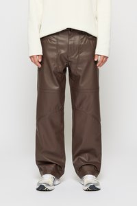 Persona vestida con pantalones anchos de imitación de cuero marrón oscuro, top blanco de manga larga y zapatillas de running blancas, de pie contra un fondo liso.