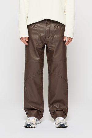 Person wearing dark brown wide-leg faux leather pants, white long-sleeve top, and white running shoes, standing against a plain background.