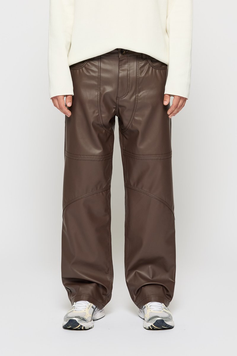 Persona vestida con pantalones anchos de imitación de cuero marrón oscuro, top blanco de manga larga y zapatillas de running blancas, de pie contra un fondo liso.