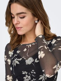 Läpinäkyvä musta paita, jossa kukkakuvio kerman ja beigen sävyissä; mallilla hopeiset kuutiokorvakorut punaisin yksityiskohdin, hiukset auki.