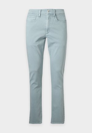 Jeune homme debout, les mains dans les poches, portant un t-shirt blanc uni, un pantalon bleu clair et des baskets blanches sur un fond blanc.