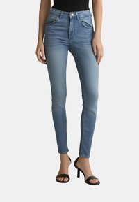 Jeans in denim blu a vita alta con un lavaggio sfumato, cinque tasche e chiusura frontale a bottoni, abbinati a sandali neri con cinturini e tacco alto.