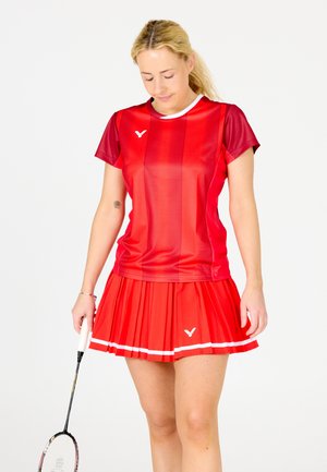 Rød sportsoutfit med en kortærmet top med lodrette striber og en plisseret nederdel. Hvide accenter og logo-detaljer på tøjet.