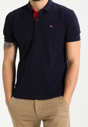 Mand iført en mørkeblå poloshirt med et lille flaglogo og beige bukser, stående med hænderne i lommerne.