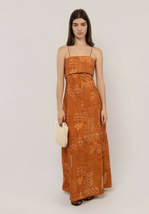 Joven mujer con un vestido maxi naranja sin mangas con estampado, sosteniendo un pequeño bolso beige, de pie contra un fondo claro y liso.