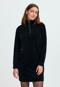 Zwarte corduroy jurk met hoge kraag en half-ritssluiting. Lange mouwen en textuurstof. Minimalistisch ontwerp, tot de knie.