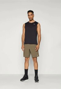 Čierny bezrukávový športový top, olivové šortky, čierne tenisky a tmavé ponožky. Výrazný detail loga na tope a šortkách. Jednoduchý, štíhly dizajn.