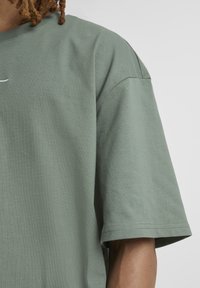 Person trägt ein locker sitzendes hellgrünes T-Shirt mit kurzen Ärmeln und sichtbaren Nähten an Schulter- und Ärmelsäumen vor einem einfarbigen Hintergrund.