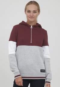Sweat-shirt à capuche bordeaux et gris avec des manches blanches, présentant une demi-fermeture éclair et des poignets côtelés. Patch logo noir en bas. Texture douce.