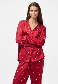 Femme avec de longs cheveux bruns portant un pyjama en satin rouge avec des motifs de cœurs blancs, se tenant les bras croisés contre un fond blanc.