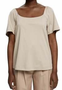 Femme portant un haut beige à manches courtes avec un large décolleté carré et un pantalon beige assorti sur fond blanc.