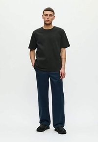 Jaunas vyras stovi su viena ranka kišenėje, vilkėdamas paprastą juodą marškinėlį, tamsiai mėlynas laisvas kelnes ir juodus batus ant balto fono.