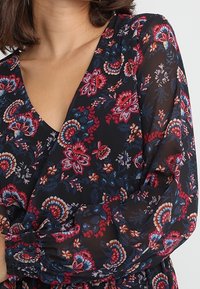 Blumenkleid aus durchsichtigem schwarzem Stoff, mit V-Ausschnitt und langen Ärmeln. Das Design umfasst rote, rosa und blaue Blumenmuster sowie geraffte Bündchen.