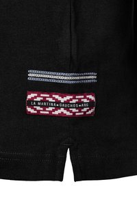 Svart tyg har en ficka med blå och silvertrådar, tillsammans med en vinröd etikett med ett geometriskt mönster och vit text som säger "La Martina Gauchos ARG."