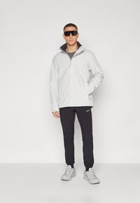 Chaqueta con cremallera de color gris claro y detalles en negro, llevada sobre una camiseta gris, combinada con pantalones negros ajustados y zapatillas grises. Gran gafas de sol oscuras.
