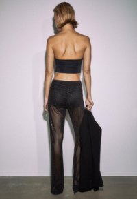 Czarny top typu bezramiączkowego zestawiony z przezroczystymi czarnymi spodniami z wzorem w kropki. Modelka trzyma czarny element odzieży, prezentując dopasowaną sylwetkę.
