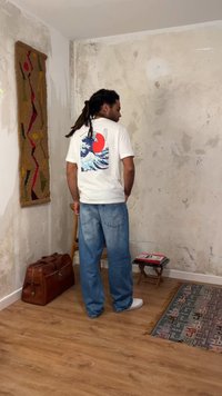 Άνδρας με dreadlocks που φοράει λευκή γραφική μπλούζα και μπλε τζιν στέκεται σε ένα δωμάτιο με υφασμένους τοίχους, ξύλινο δάπεδο, υφαντή τοιχογραφία και δερμάτινη τσάντα.