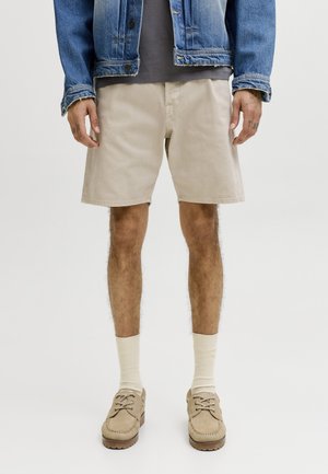 Persoană purtând o jachetă de blugi albastră, un tricou gri, pantaloni scurți bej, șosete crem și mocasini bej, stând în fața unui fundal simplu.