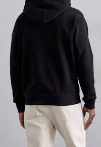 Sudadera negra con capucha hecha de tela suave, con un bolsillo tipo canguro y puños acanalados, combinada con pantalones de color claro.
