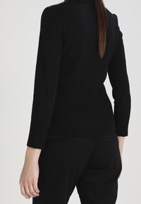 Femme vue de dos portant un pull ajusté noir à manches longues et un pantalon noir avec des cheveux bruns raides tombant.