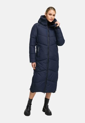 Woman wearing a long navy blue padded winter coat with a hood, front zipper, and black ankle boots against white background.