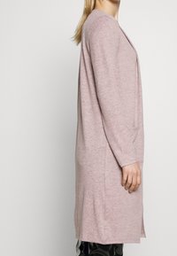 Lång, ljusrosa kofta i en mjuk stickad textur, med smal passform, långa ärmar och sidofickor vid fållen för bättre rörlighet.