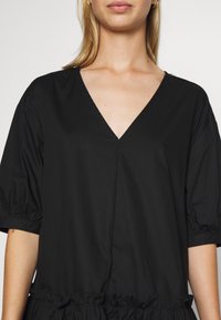 Černé šaty s výstřihem do V, krátkými nabíranými rukávy a skládacími volány na lemu. Vyrobené z hladkého, lehkého materiálu.