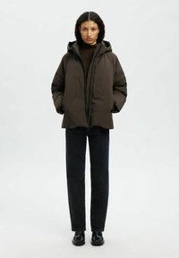 Giacca marrone imbottita con cappuccio, chiusura con zip e polsini elastici, abbinata a jeans in denim scuro e scarpe nere. Design semplice e funzionale.