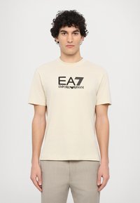 béžové bavlněné tričko s krátkým rukávem, na přední straně s černým logem EA7 Emporio Armani. Jednoduchý design s kulatým výstřihem.