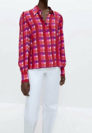 Blusa a maniche lunghe con un motivo a quadri rosso e rosa, con spalle a puffo e chiusura a bottoni, abbinata a pantaloni bianchi.