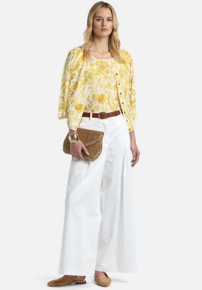 Femme portant un chemisier jaune à fleurs et un cardigan, un pantalon blanc à jambes larges, une ceinture marron, tenant une pochette tissée et portant des ballerines tissées.