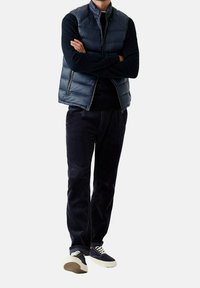 Marineblaue Steppweste über einem dunklen Langarmshirt, kombiniert mit dunkler Cordhose und marineblauen Sneakers mit weißen Schnürsenkeln und Sohlenakzenten.