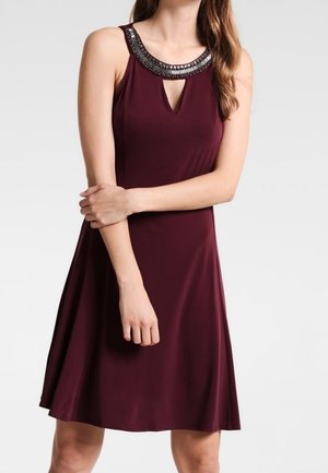 Donna che indossa un vestito bordeaux senza maniche con dettaglio di perline sullo scollo e apertura a goccia sul davanti, in piedi con le braccia incrociate.