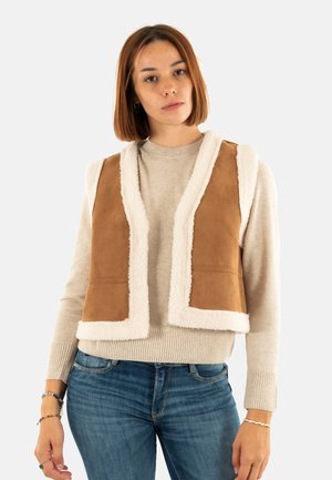 Bruine faux-suède gilet met een fluffy witte voering, met een open voorkant, zonder sluitingen en twee zijzakken. Draagt over een lichte trui.