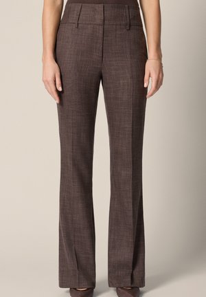 Kobieta nosząca brązowe, wysokie taliowane spodnie z przednią kantą, prostymi nogawkami i bocznymi kieszeniami, stojąca na neutralnym tle.