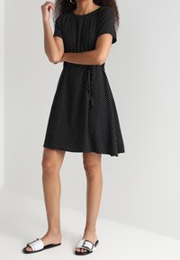 Robe noire à pois avec des manches courtes et une taille à cordon, fabriquée en tissu léger. Modèle portant des sandales à glisser noires et blanches.