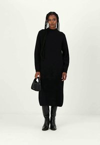 Femeie cu părul împletit în bretele lungi, îmbrăcată într-o rochie midi neagră cu mâneci lungi, purtând cizme negre și ținând o geantă mică neagră pe un fundal deschis.