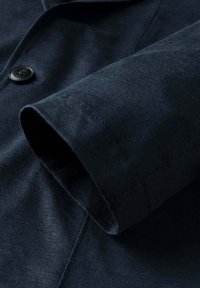 Detailný záber na rukáv tmavomodrého saka s viditeľným prešívaním a čiernym gombíkom na prednej časti.