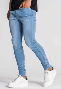 Jean stretch bleu clair avec une coupe slim, comportant un design à cinq poches et des coutures discrètes. Portés avec des baskets blanches avec un accent noir.