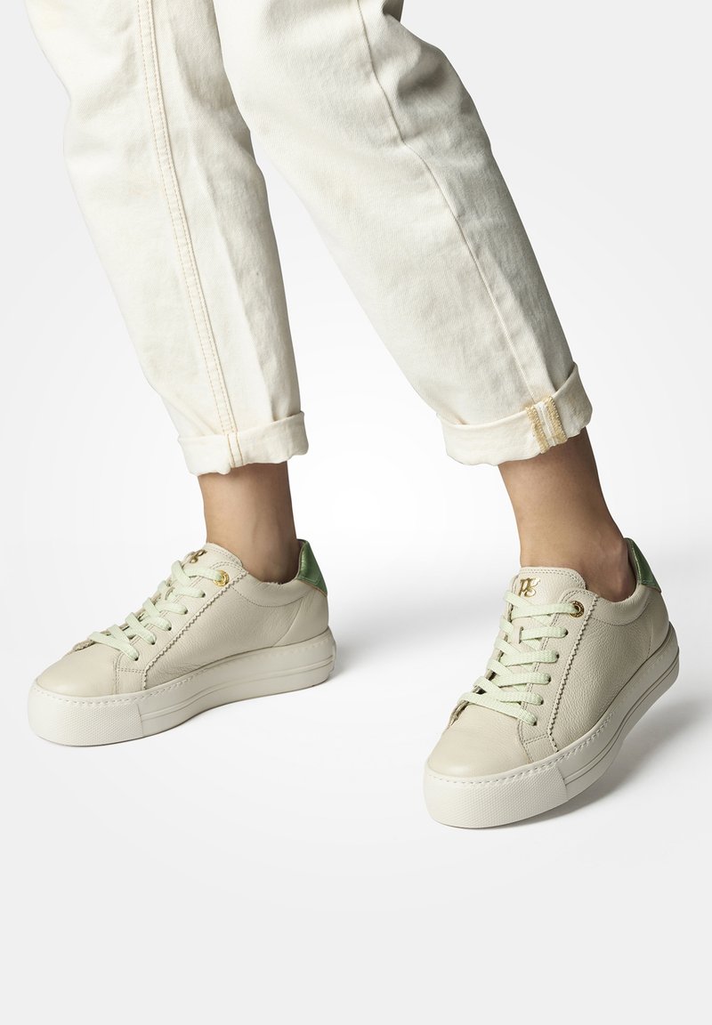 Sneakers in pelle beige con finitura testurizzata, caratterizzati da un'accenno di tallone verde chiaro e lacci sottili, abbinati a pantaloni bianchi a orlo.
