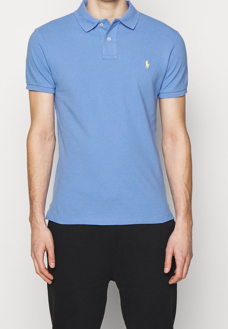 Homme portant un polo bleu clair à manches courtes avec un petit logo jaune sur la poitrine et un pantalon noir, debout devant un fond uni.