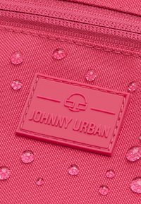 Piros vászonzsák, amelynek címkéjén a "JOHNNY URBAN" felirat található, fényes cseppekkel és texturált felülettel. Egy sima, rózsaszín logóval díszítve.