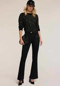 Jeans neri con gamba a zampa abbinati a un maglione di maglia a strisce zebra nero e verde, in abbinamento a un cappello nero e a una borsa lavorata.