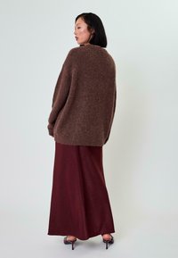 Rjav puhast oversized pulover, kombiniran z dolgo bordo krilo, ki ima gladko teksturo in diskretne detajle na robu.